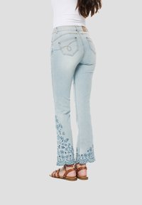Svetlomodré džínsy s kvetinovou výšivkou na lemoch, rovný strih nohavíc a jemne vyblednutý vzhľad. Viditeľné hnedé sandále s remienkami.
