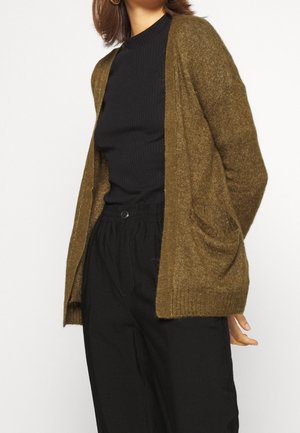 Kobieta ubrana w oliwkowozielony, otwarty sweter dziany na przód, czarny prążkowany top i luźne czarne spodnie, z złotymi kolczykami kołami.