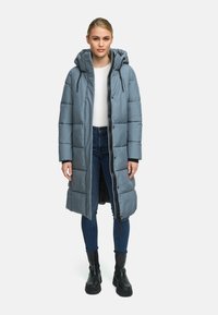 Manteau long rembourré en bleu clair, avec une capuche, une fermeture éclair frontale et des poches latérales. Porté sur un haut blanc et un jean avec des bottes noires.