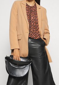 Svart handväska med krokodilstruktur som hålls av en person klädd i en rosa blazer över en mönstrad topp och byxor i konstläder.