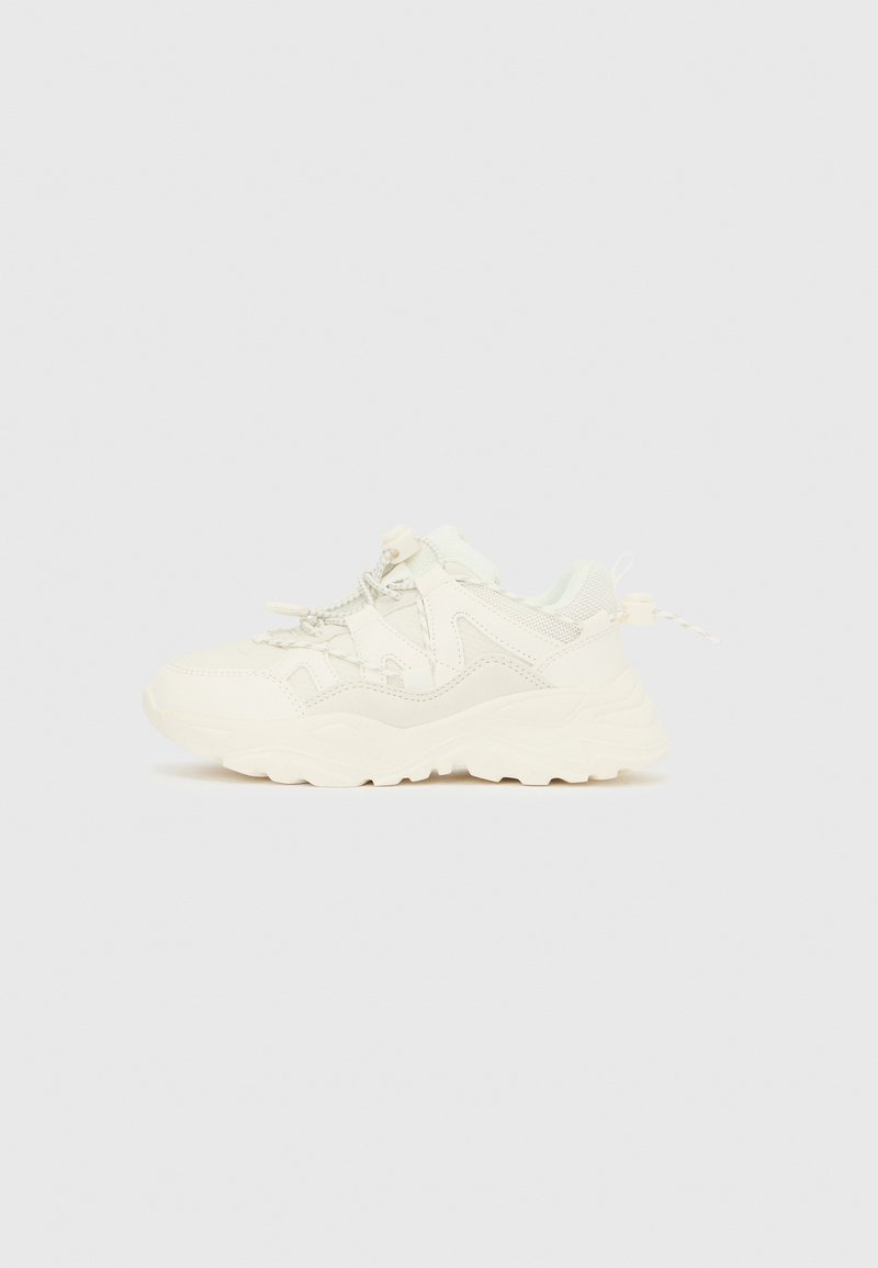 Fehér, vastag talpú sportcipő hálós és bőrpanelekkel, vastag talppal, valamint bungee stílusú fűzőkkel, állítható csattal.