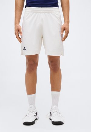 Uomo che indossa pantaloncini sportivi bianchi Adidas, calzini bianchi e scarpe da ginnastica Adidas bianche, in piedi contro uno sfondo chiaro uniforme.