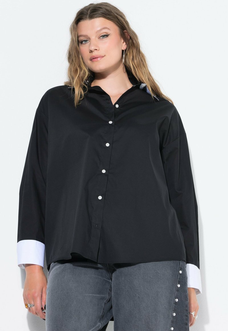 Giovane donna con capelli biondi mossi che indossa una camicia nera con bottoni e polsini bianchi, e jeans grigi con bottoni laterali su uno sfondo bianco.