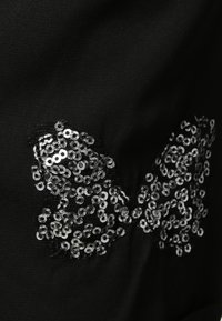 Tissu noir présentant une forme de papillon en paillettes argentées, détaillée avec un contour en mesh et une surface texturée.