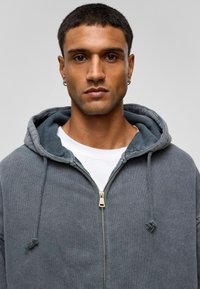 Grijs zip-up hoodie met een zachte textuur, voorzien van een gouden rits, trekkoorden en laag over een wit T-shirt. De capuchon is gevoerd met een donkere tint.