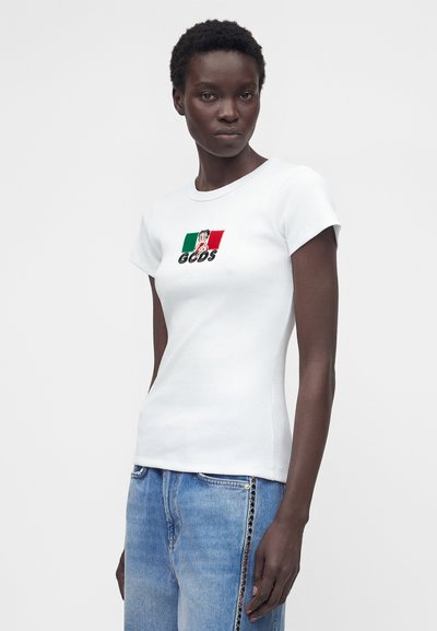 Γυναίκα που φοράει εφαρμοστό λευκό μπλουζάκι με πράσινο, λευκό και κόκκινο λογότυπο και ανοιχτό μπλε τζιν με κουμπιά στο πλάι.