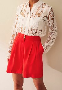 Witte kanten blouse met bloemenpatronen, lange mouwen en een knoopsluiting bij de kraag. Rode geplooide shorts met zijzakken en twee knopen aan de voorkant.