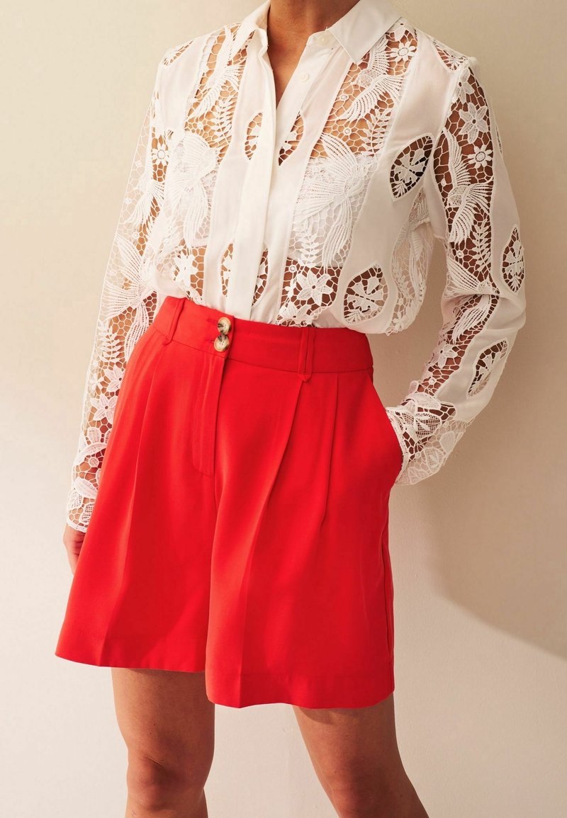 Witte kanten blouse met bloemenpatronen, lange mouwen en een knoopsluiting bij de kraag. Rode geplooide shorts met zijzakken en twee knopen aan de voorkant.