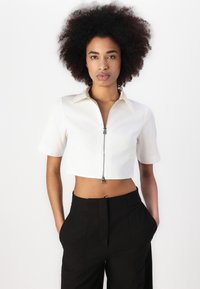 Camisa blanca corta con mangas cortas y cremallera en la parte frontal, combinada con pantalones negros de cintura alta. Tejido suave, diseño minimalista.