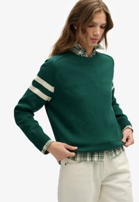 Zielony sweter z dzianiny z białymi paskami na rękawach, o teksturowanym wykończeniu. Noszony na koszuli w kratę. Luźny krój z prążkowanymi mankietami i dołem.