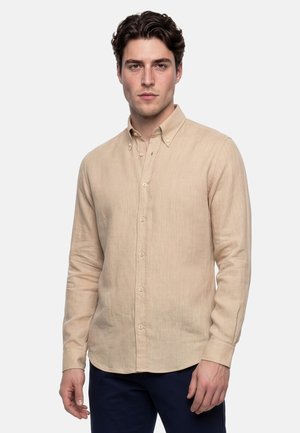 Jeune homme portant une chemise beige à manches longues boutonnée et un pantalon foncé, debout devant un fond clair uni.