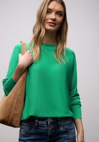 Jeune femme portant un pull vert et un jean, tenant un grand sac à bandoulière en daim beige clair sur un fond gris clair uni.