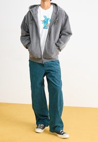Grauer Kapuzenpullover mit Frontreißverschluss, weißes T-Shirt mit einem blauen Hasenmotiv und türkisfarbene Cordhosen. Schwarze Sneakers runden das Outfit ab.