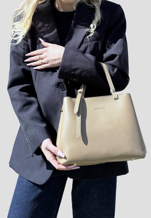 Donna con capelli biondi lunghi che indossa un blazer nero a righe tiene in mano una borsa in pelle beige con ferrature dorate.