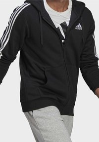 Sweat à capuche zippé noir en tissu avec des accents blancs des trois bandes Adidas sur les manches. Comprend une capuche avec cordon de serrage et des poches avant.