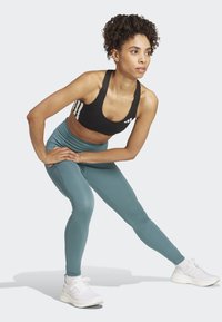 Reggiseno sportivo nero con strisce bianche, leggings turchesi e sneakers bianche. La figura è in una posa di stretching, evidenziando l'adattamento e il design dell'outfit.