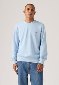 Φόρμα λαιμόκοψης σε ανοιχτό γαλάζιο, από βαμβακερό μείγμα, με ριμπ μανσέτες και τελείωμα, με ένα μικρό κόκκινο λογότυπο στο στήθος. Φοράγεται με μπλε τζιν.