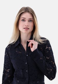 Camicia azzurra in pizzo con bottoni, caratterizzata da motivi floreali, colletto e design fitted con una leggera lucentezza e una finitura texture.