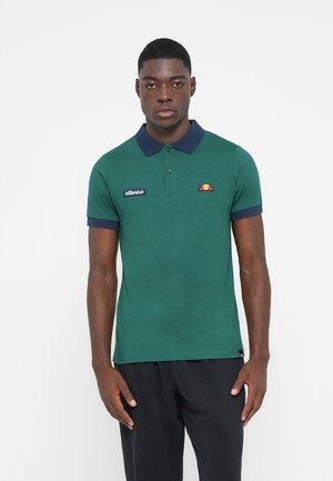 Grønn polo skjorte laget av bomull, med marinefarget krage og mansjetter, et brodert logo på venstre bryst, og et lite fargerikt merke.