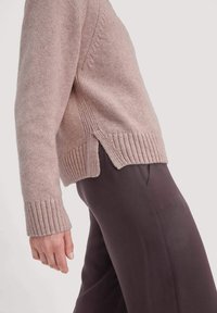 Person, der einen hellpink gestrickten Pullover mit seitlichem Schlitz und dunklen braunen Hosen vor einem einfarbigen Hintergrund trägt.