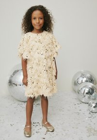 Rochie bej cu paiete, design cu franjuri texturat și mâneci scurte. Purtați cu pantofi Mary Jane aurii, stând lângă mingi disco argintii.