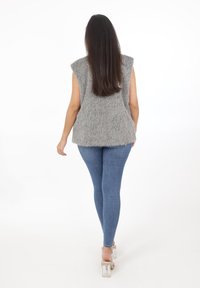 Szary bezrękawnik z sztucznego futra o luźnym kroju, z teksturowaną powierzchnią, zestawiony z niebieskimi dżinsami slim-fit i przezroczystymi sandałami na wysokim obcasie.