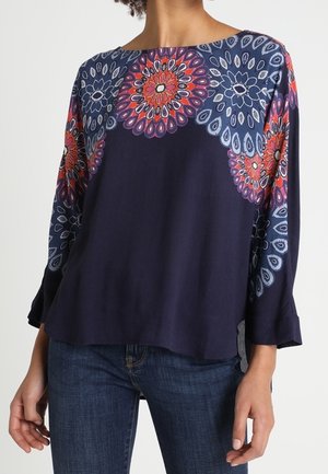 Laivastonsininen pitkähihaisen paita, jossa punaiset ja siniset pyöreät kukkakuviot hartioissa ja hihojen yläosassa, yhdistetty tummansinisiin farkkuihin.