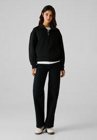 Sudadera negra de pullover con cuello medio abotonado, llevada sobre una camisa blanca, combinada con jeans oscuros de pierna recta y zapatillas blancas.