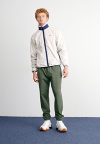 Chaqueta de cremallera color beige claro con acentos en azul marino y un patrón intrincado, combinada con pantalones verdes y zapatillas blancas con suelas marrones.