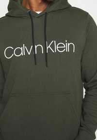 Felpa con cappuccio verde oliva scuro in tessuto morbido. Presenta un logo "Calvin Klein" bianco, una tasca frontale e stringhe nere con punte in metallo.