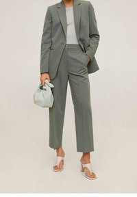 Femme portant un blazer vert sauge et un pantalon large, avec un haut blanc, portant un petit sac à main menthe et des sandales à talons ouverts blanches.