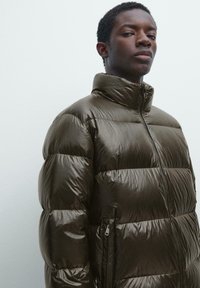 Steppjacke in dunklem Olivgrün mit hohem Kragen, gestepptem Design, durchgehendem Reißverschluss vorne und Reißverschlusstaschen an den Seiten. Glänzendes Material.
