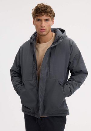 Un hombre luce una chaqueta con capucha de color gris oscuro sobre un jersey beige y posa con las manos en los bolsillos sobre un fondo liso y claro.