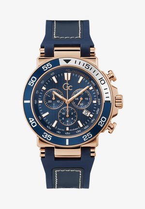 Zegarek na rękę w kolorze różowego złota z niebieskim paskiem z gumy. Posiada niebieską bezel tachymetryczną, fakturowaną ciemnoniebieską tarczę, trzy małe tarcze oraz okno daty.