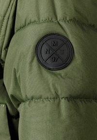 Section d'une veste en doudoune verte avec une texture matelassée, des coutures visibles et un badge en caoutchouc noir rond portant le logo "MNT 97".