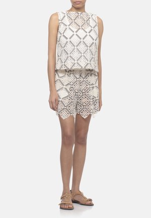 Donna che indossa un top in pizzo bianco senza maniche e pantaloncini abbinati con motivo geometrico, abbinati a sandali aperti color cuoio, in piedi su sfondo bianco.