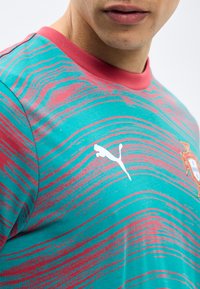 Suurkava inimene, kes kannab türkiissinist ja roosat mustrilist spordipulka, millel on Puma logo ja osaline sporditegaemblem rinnal.