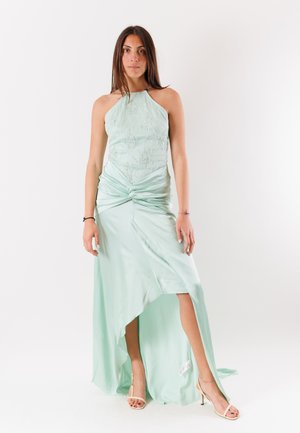 Donna in piedi con un abito verde chiaro a collo halter con ricamo floreale e uno spacco alto, indossando sandali beige con tacchi alti.