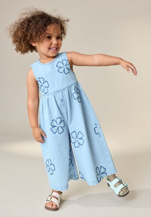 Peuter met krullend haar die een mouwloze brede jumpsuit met blauwe bloemenprint en witte sandalen draagt, glimlacht en haar arm uitstrekt.