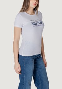 T-shirt bianca in cotone con collo rotondo, caratterizzata da un ricamo audace blu con il logo "EAA" e "Emporio Armani", abbinata a jeans in denim azzurri.