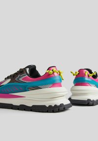 Masivní tenisky v růžové, modré a šedé barvě s černou podrážkou a žlutými tkaničkami, zobrazené z rear a bočního úhlu na šedém pozadí.