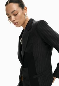 Blazer a righe nere con vestibilità sartoriale, caratterizzato da pannelli a contrasto, texture in satin e chiusura frontale con un solo bottone.