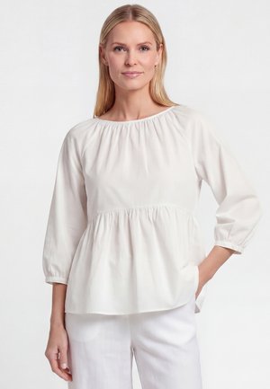 Blonde vrouw met steil haar draagt een losse witte blouse met lange mouwen en een witte broek, staand tegen een effen witte achtergrond.
