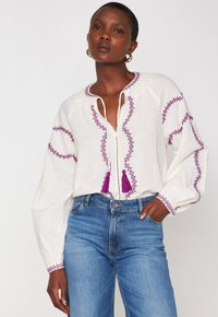Weiße bestickte Bluse mit lila Stickerei und Quasten am Ausschnitt, kombiniert mit hochgeschnittenen blauen Jeans. Lange, luftige Ärmel.