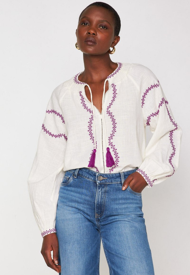 Weiße bestickte Bluse mit lila Stickerei und Quasten am Ausschnitt, kombiniert mit hochgeschnittenen blauen Jeans. Lange, luftige Ärmel.
