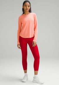Tricou sport cu mâneci lungi de culoare roz deschis, asortat cu colanți roșii fit. Ținuta prezintă materiale fine și o textură moale, ușoară.