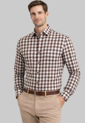 Homme portant une chemise à manches longues à carreaux marron et blanc rentrée dans un pantalon beige avec une ceinture marron, debout devant un fond gris.