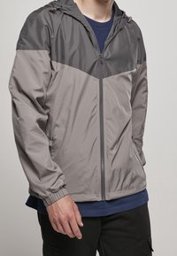 Man i grå och mörkgrå zip-up vindjacka över en marinblå skjorta, med svarta byxor, stående mot en neutral bakgrund.