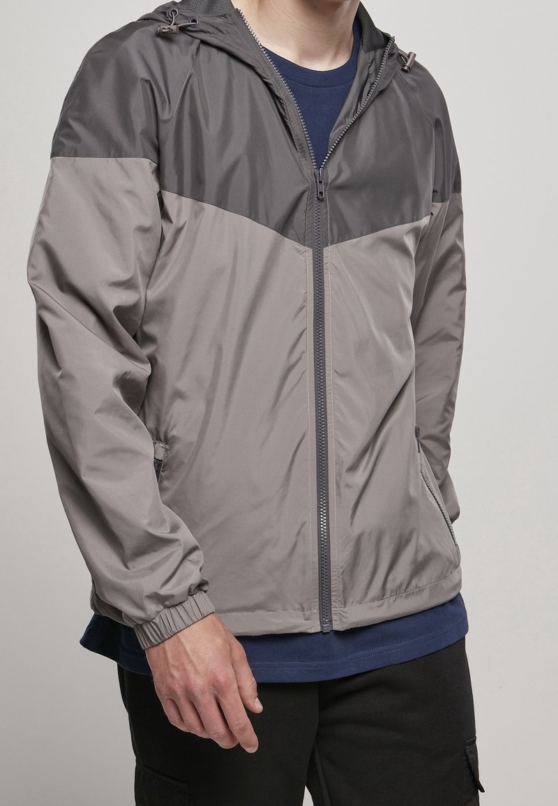 Man i grå och mörkgrå zip-up vindjacka över en marinblå skjorta, med svarta byxor, stående mot en neutral bakgrund.