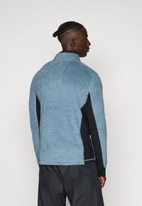Jachetă din fleece de culoare albastru deschis, cu o suprafață texturată, guler înalt și panouri laterale negre. Are un croi lejer și un material moale.